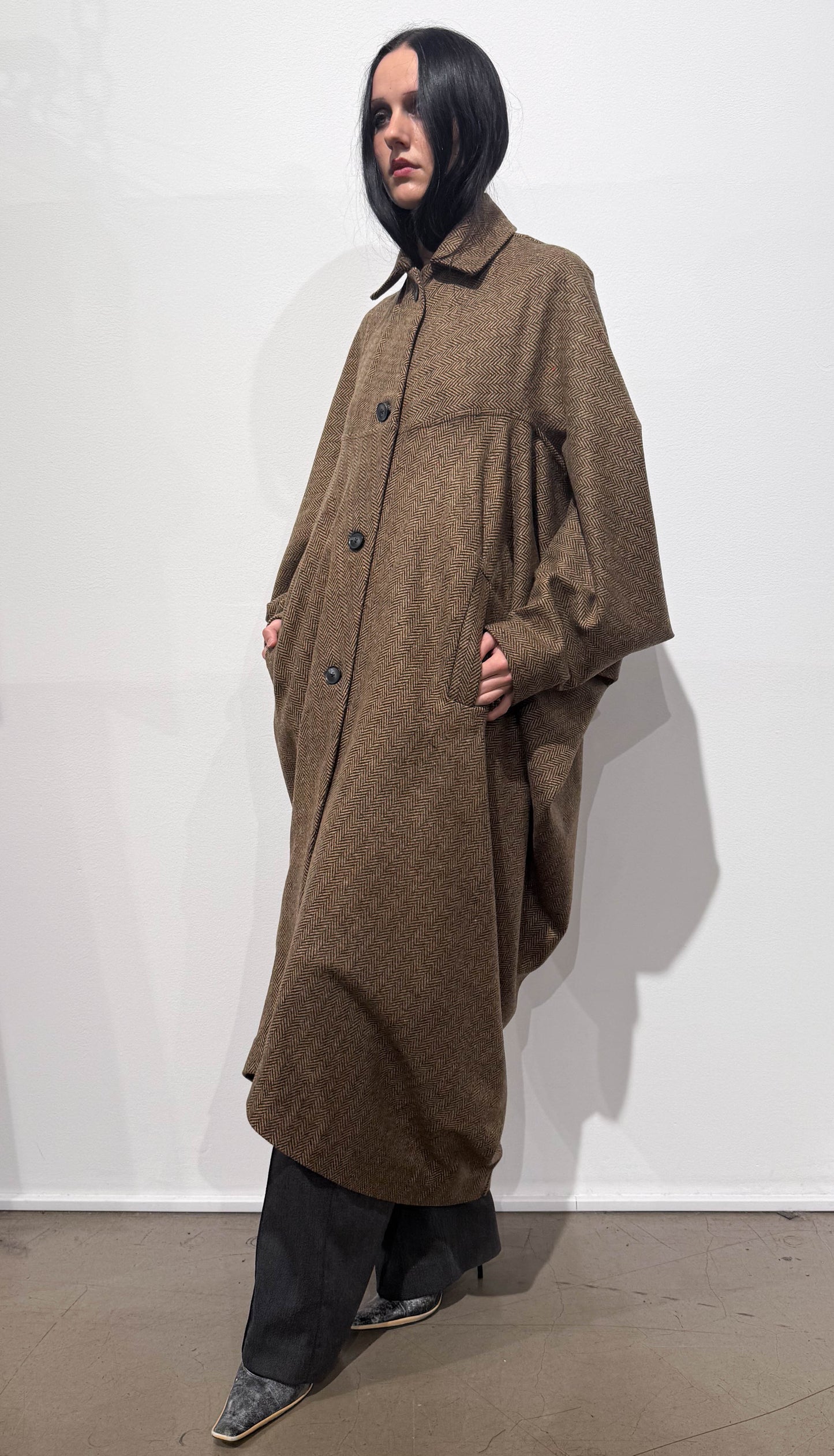 Person wearing a long brown coat against a white wall
