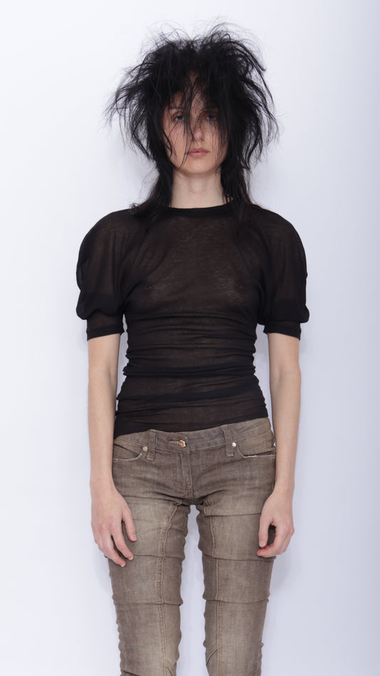 Person wearing a sheer black top and brown pants against a white background