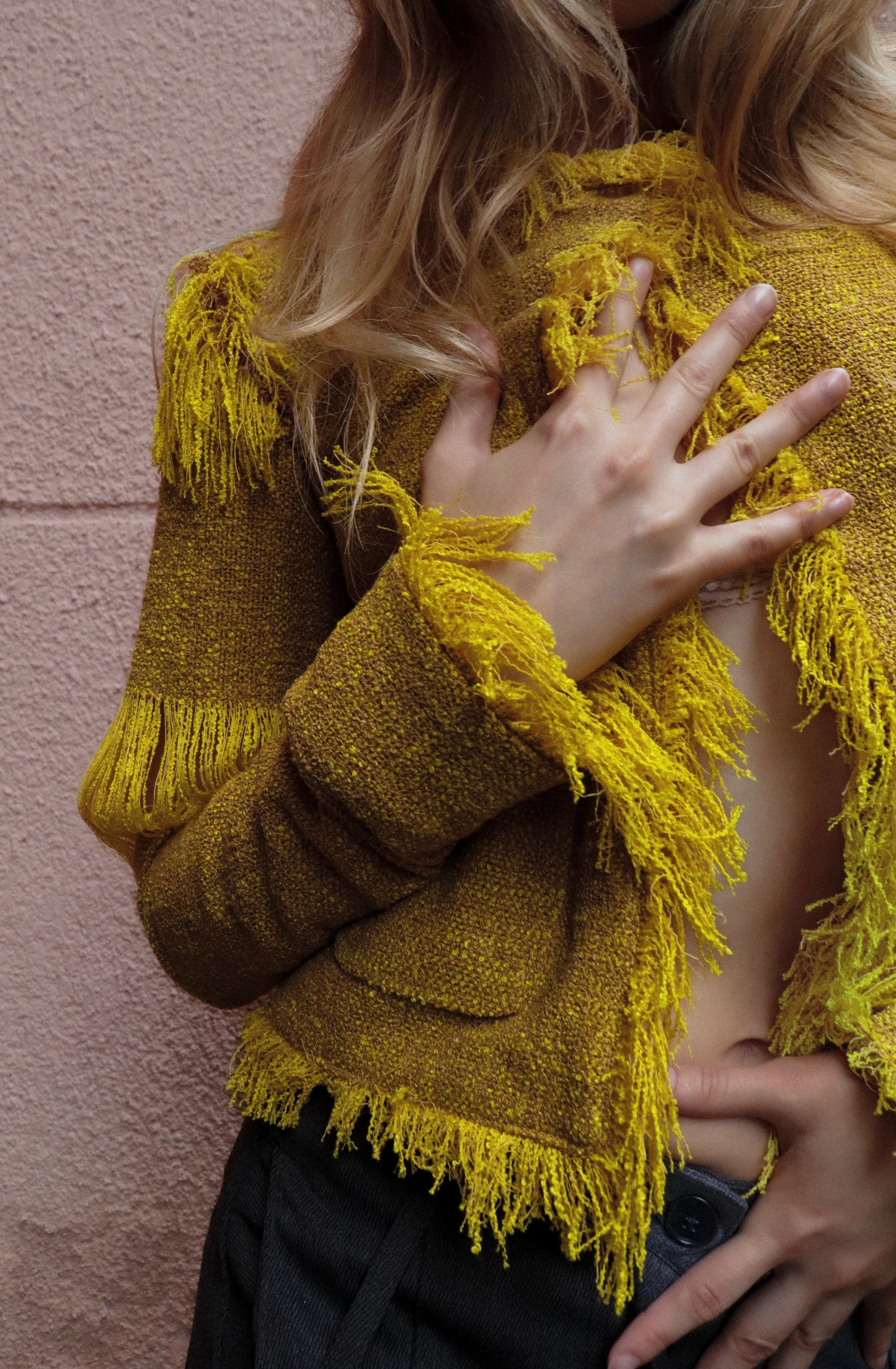 Person wearing a mustard yellow fringed jacket against a pink wall