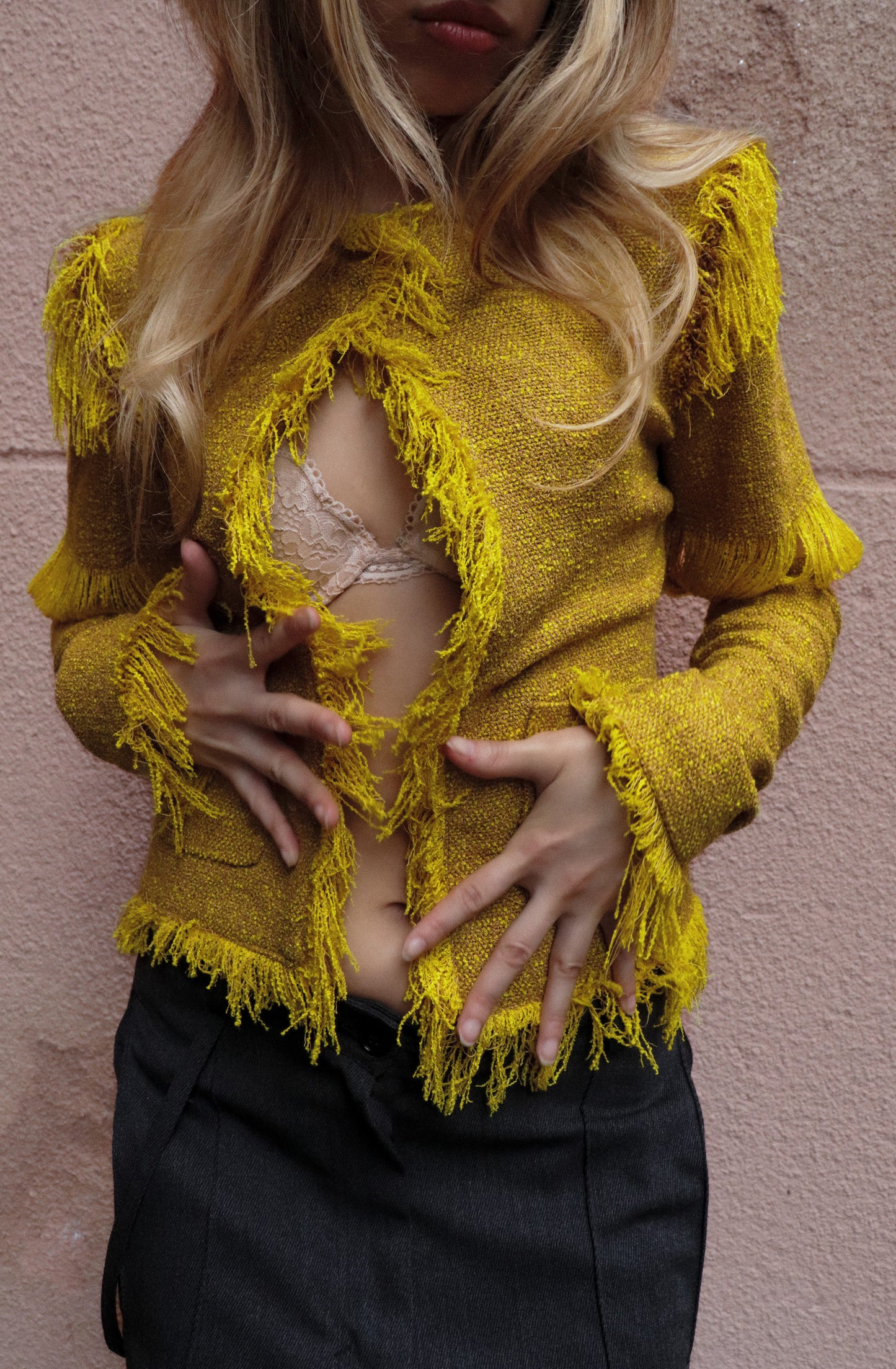 Person wearing a yellow tweed jacket over a black outfit against a pink wall.