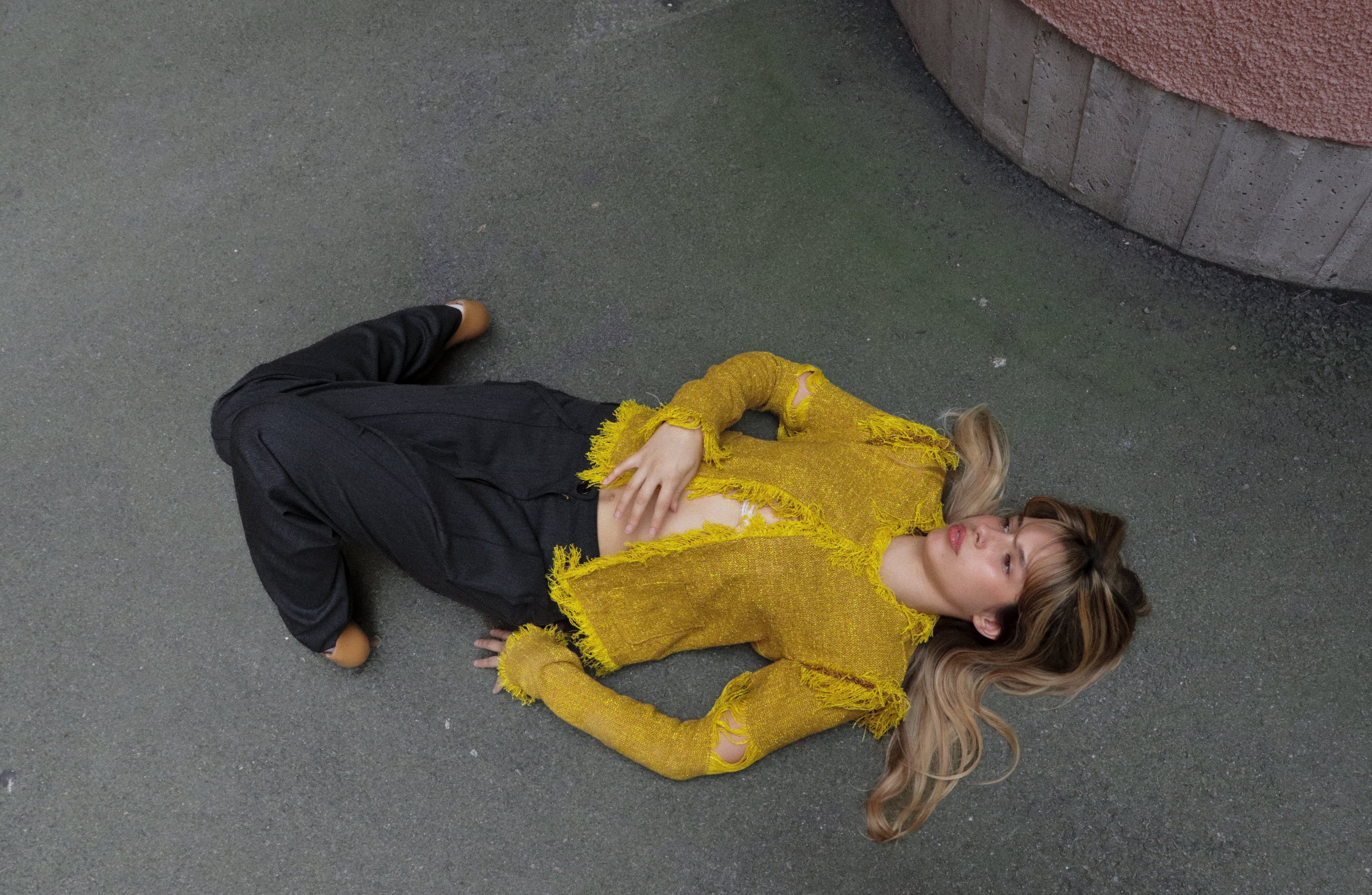 Person wearing a yellow textured top and black pants lying on a concrete floor.